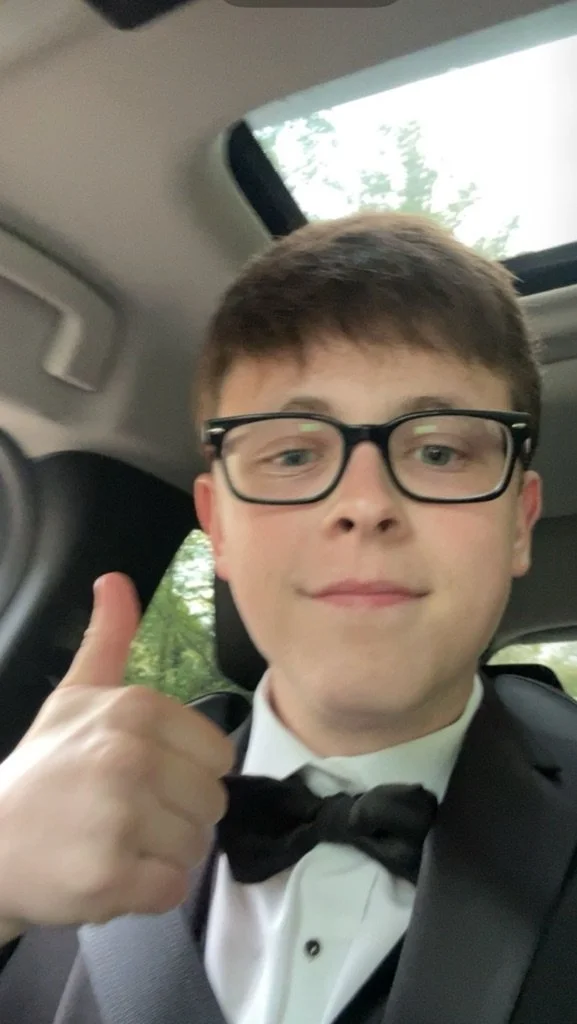 A young man with glasses in a tuxedo taking a selfie inside a car, giving a thumbs-up gesture with a tree-lined background outside.