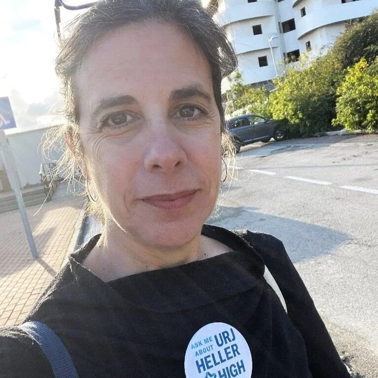 A woman in a black shirt with a button that reads "Ask me about URJ Heller High," standing outdoors in a parking lot with buildings and greenery in the background.