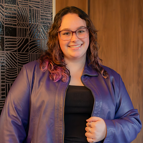 A woman with curly brown hair, wearing a black top and purple jacket, smiling.