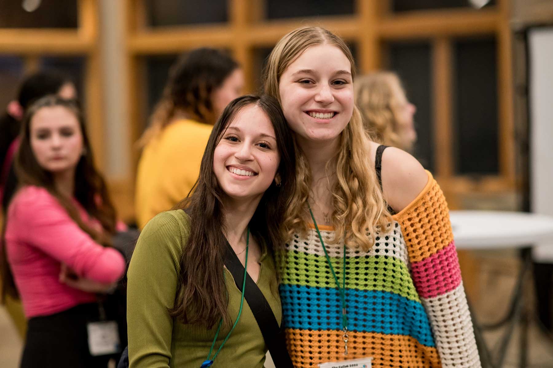 Two young women smiling at the camera at a social event, one wearing a green top and the other a colorful striped sweater.