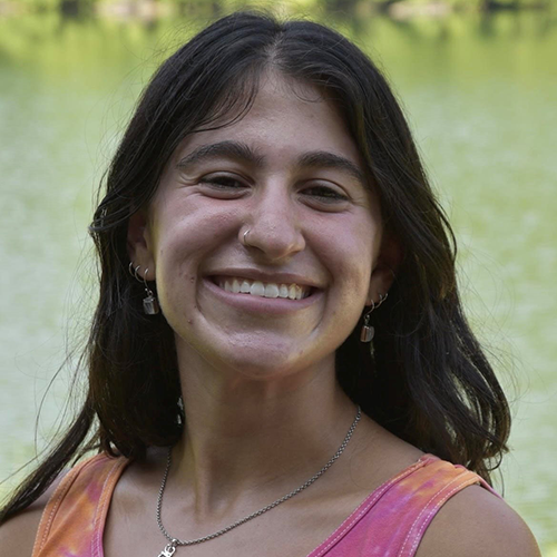 A woman with dark hair, wearing a pink shirt and a necklace, smiling and standing outdoors.