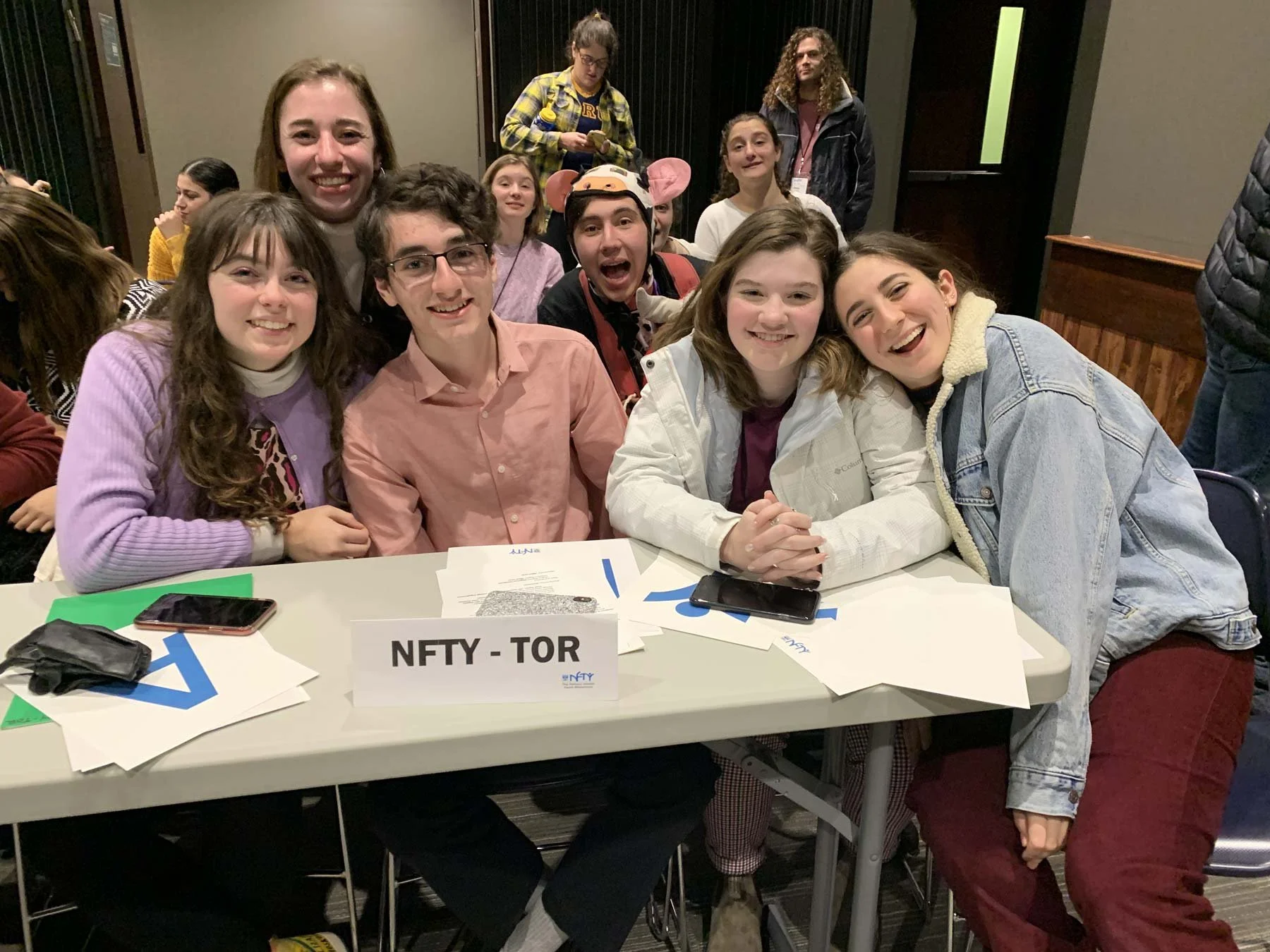 A group of young people sitting around a table at an event, with some smiling at the camera. There is a sign on the table that says 'NFTY - TOR', along with papers, a phone, and a wallet. In the background, more people are standing or sitting, and decorations are visible.