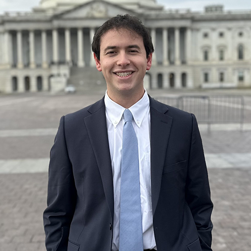 A man wearing a dark suit with a blue tie and white shirt, smiling and standing in front of the Capital Building.
