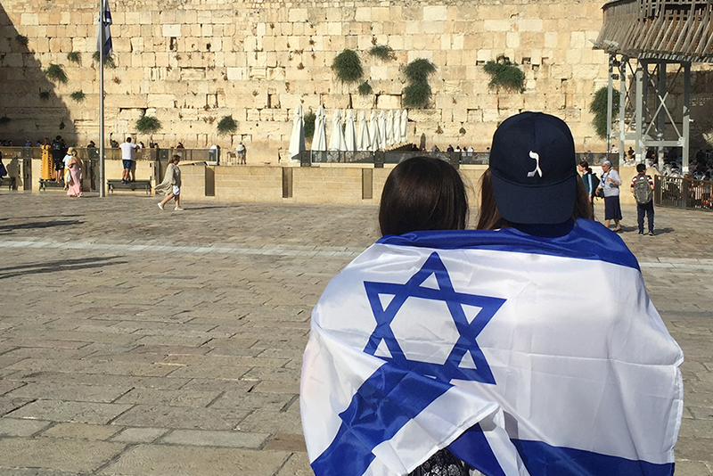 Two people sitting together in front of the Western Wall, with one person draped in an Israeli flag.