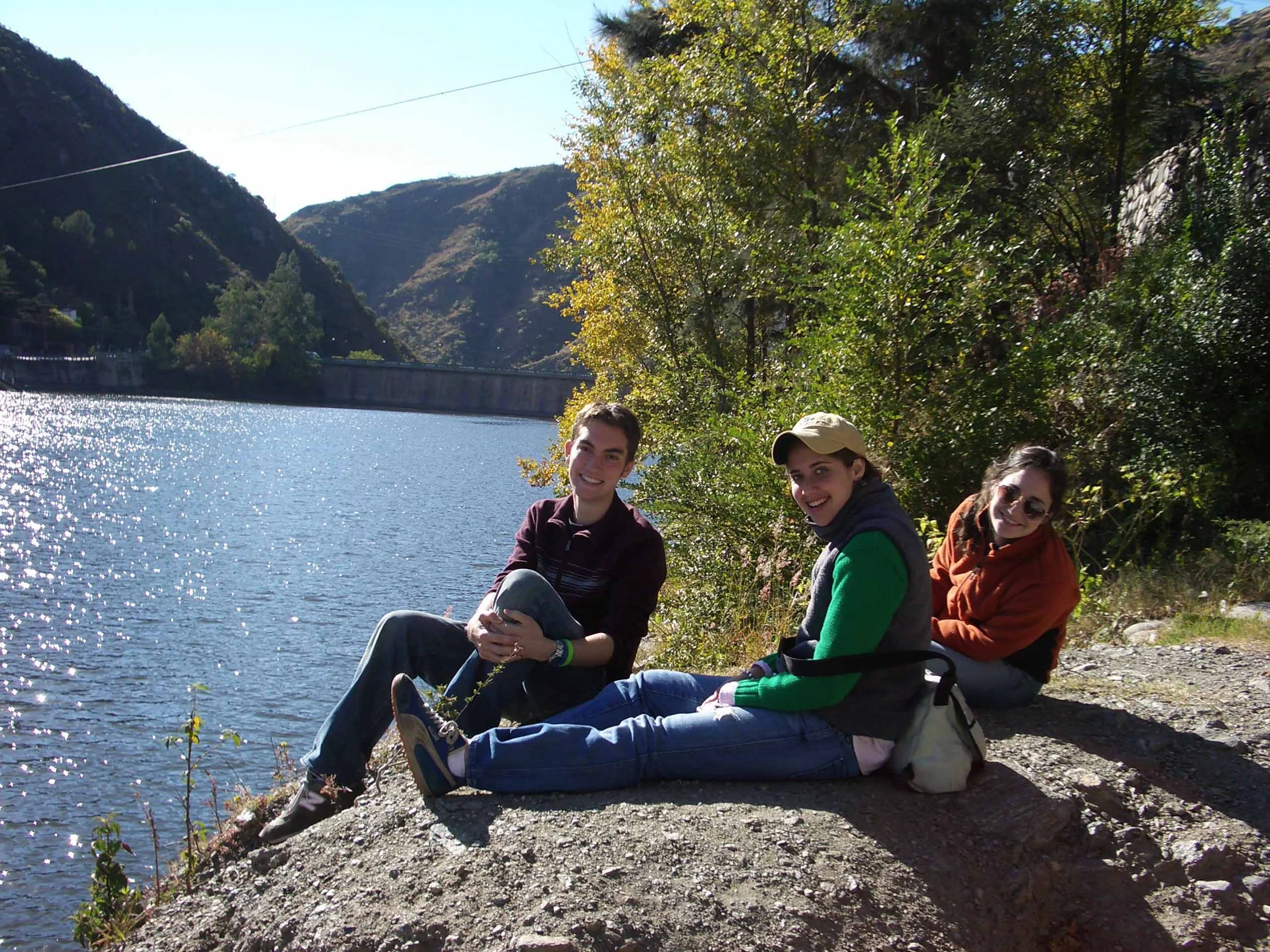 Group of young people sitting on a rocky riverbank surrounded by trees with mountains in the background, enjoying a sunny day.