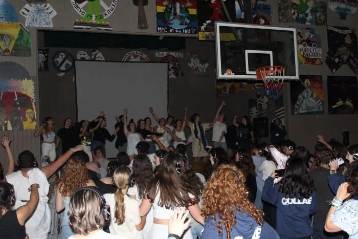 Crowd of students watching a performance on stage in a gymnasium with basketball hoop, colorful posters, and a large screen in the background.