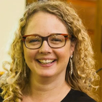 A woman with curly blonde hair wearing glasses and earrings, smiling at the camera indoors.