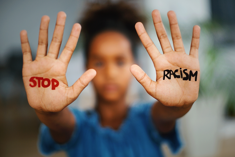 A young girl with short curly hair holding up her hands with words written on her palms: 'STOP' on the left hand and 'RACISM' on the right hand, in focus. The background is blurred.