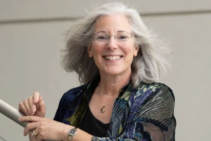 A smiling elderly woman with gray hair, wearing glasses, a colorful scarf, and a black top, holding a white object in her hands.