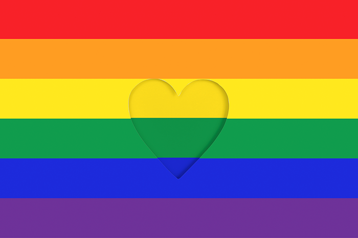 Rainbow flag with red, orange, yellow, green, blue, and purple stripes. A heart shape is cut out in the center of the yellow and green stripes.