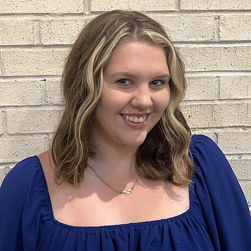 A woman with wavy blonde hair wearing a blue top, smiling and standing in front of a white brick wall.