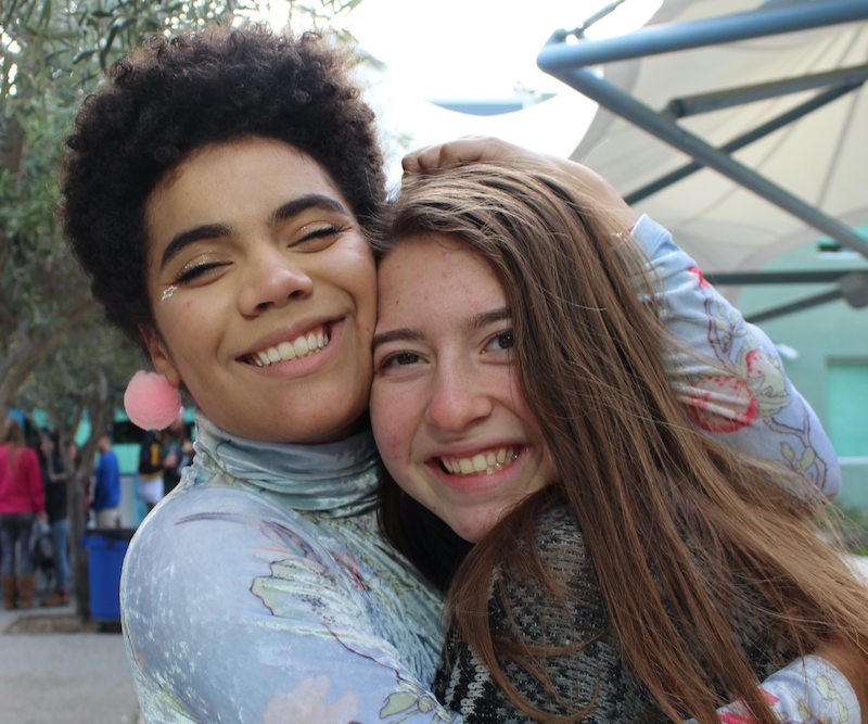 Two young people hugging and smiling outdoors at a park or fair, with trees, other people, and a canopy in the background.