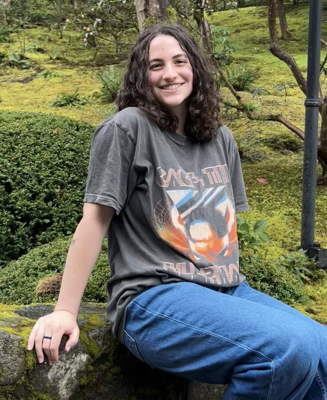A woman with curly brown hair smiling and sitting on a moss-covered rock in a lush green garden or park, wearing a gray graphic t-shirt and blue jeans.