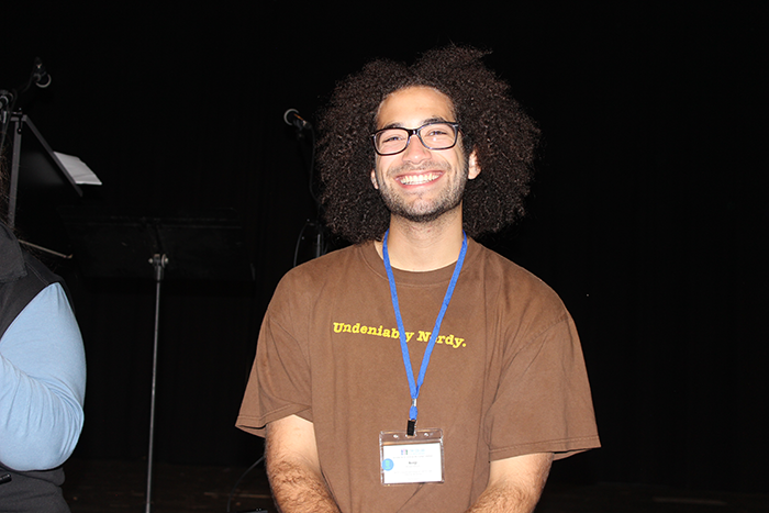 A man with curly hair, glasses, and a beard smiling, wearing a brown t-shirt with yellow text and a blue lanyard with an ID badge.