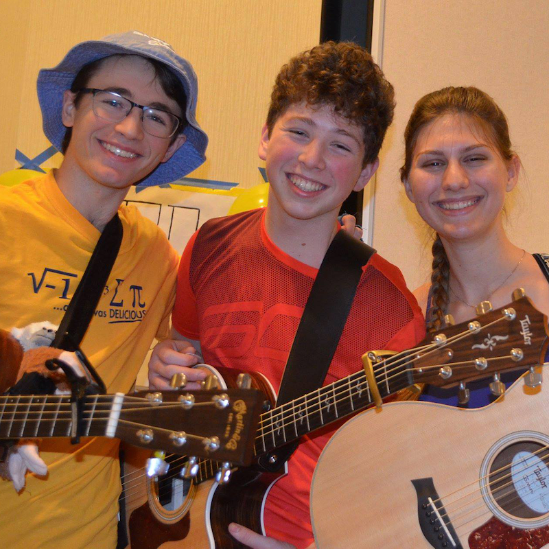 Three young adults smiling and holding guitars at an indoor event.
