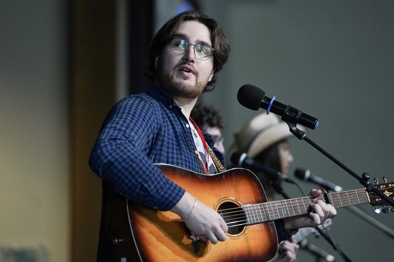 A man with glasses, a beard, and long hair playing an acoustic guitar and singing into a microphone during a performance.