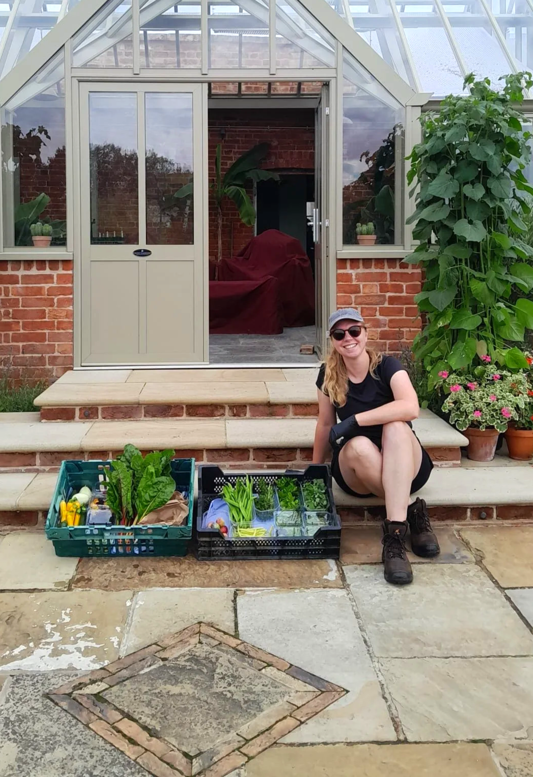 A picture of Emily, the Gloucestershire Gardener, sat, smiling on some glasshouse steps, next to two crates of harvested vegetables.