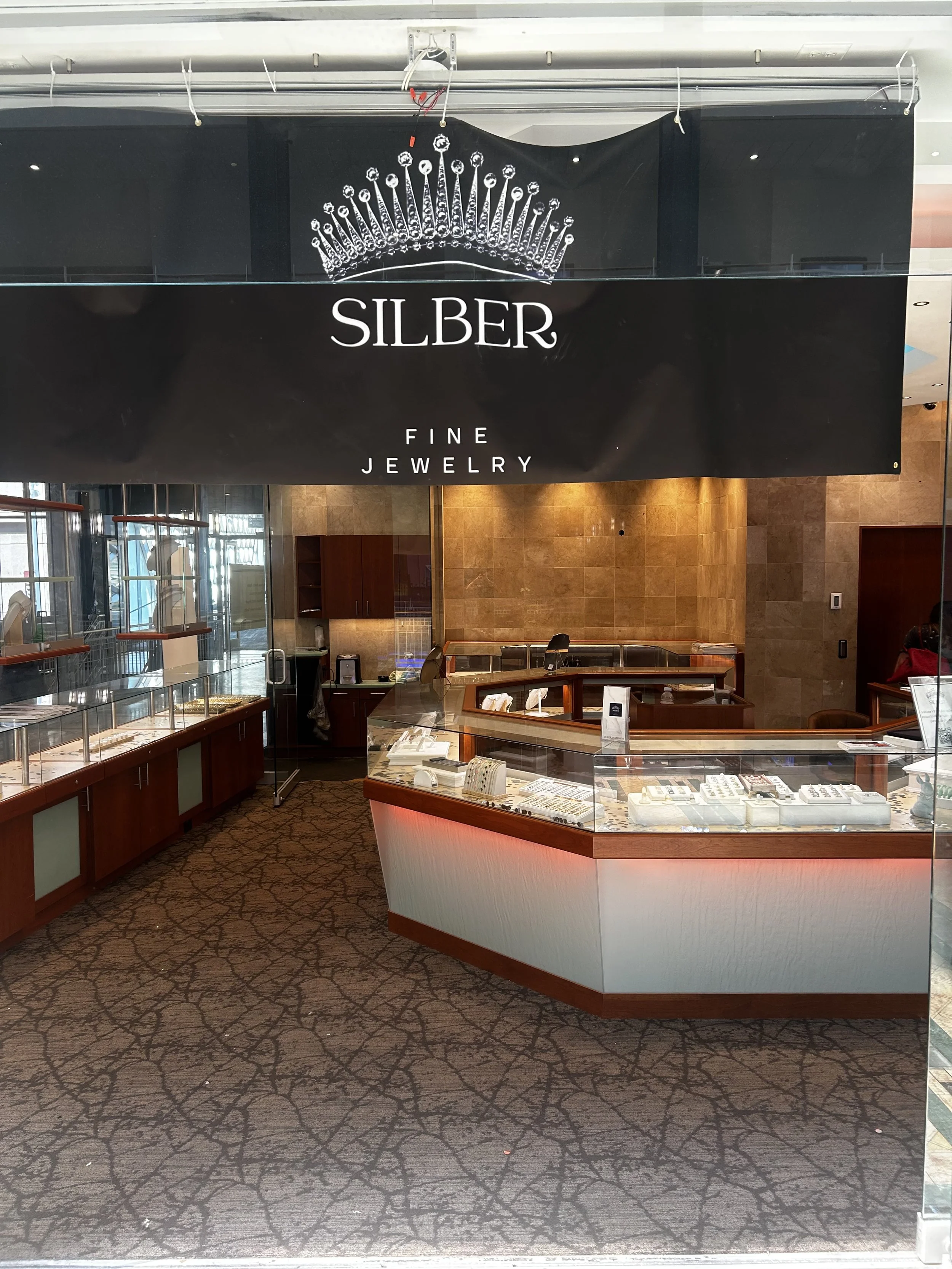 Jewelry store display with glass cases showing rings and necklaces, and a sign that reads 'Silber Fine Jewelry' with a crown logo above.
