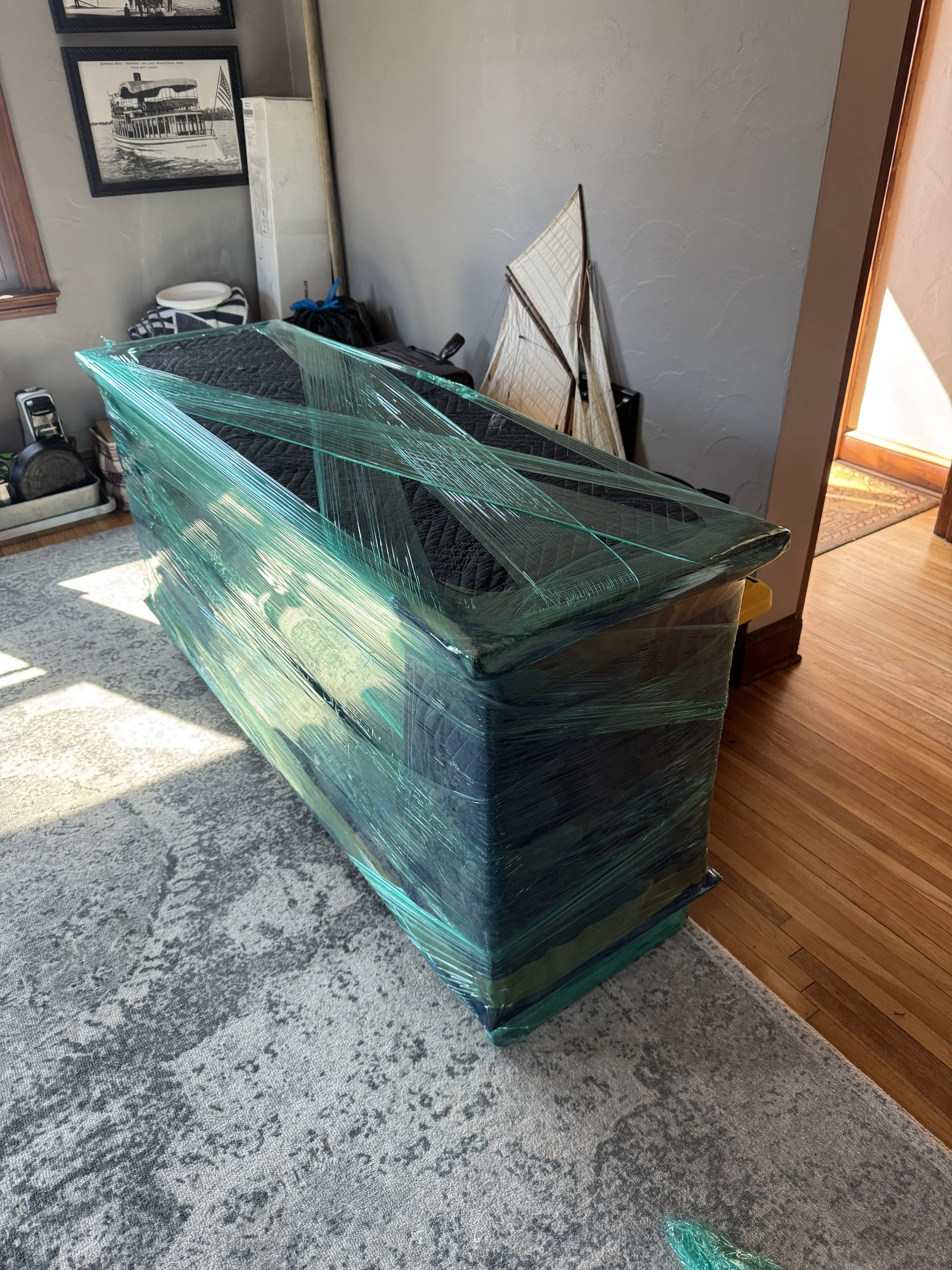 A wrapped large object, possibly a sofa or furniture piece, with green plastic wrapping in a living room. There's a rug, framed pictures, and various household items visible in the background.