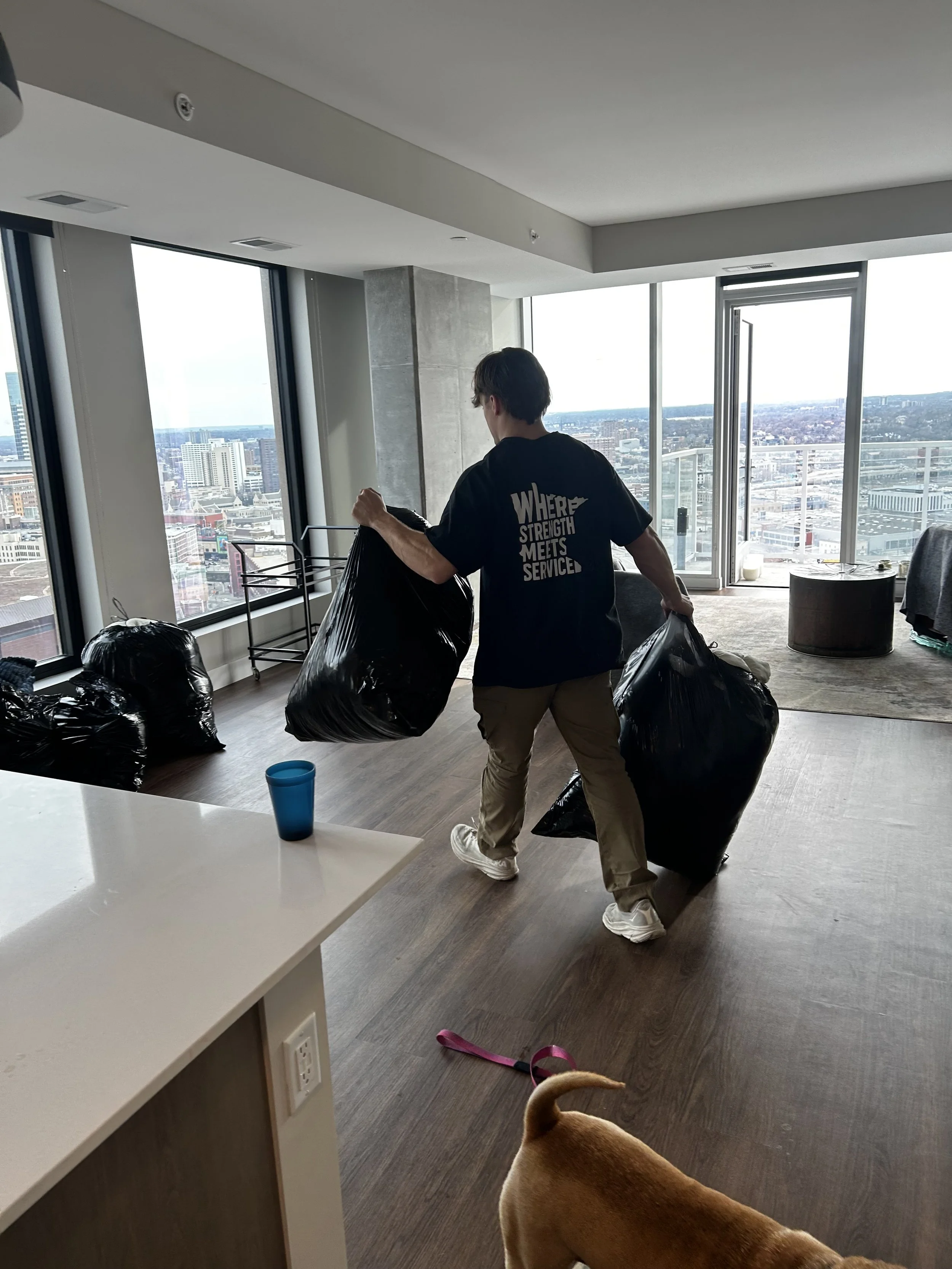 A man carrying large black trash bags inside a modern apartment with large windows and a city view, with a dog partially visible in the foreground.