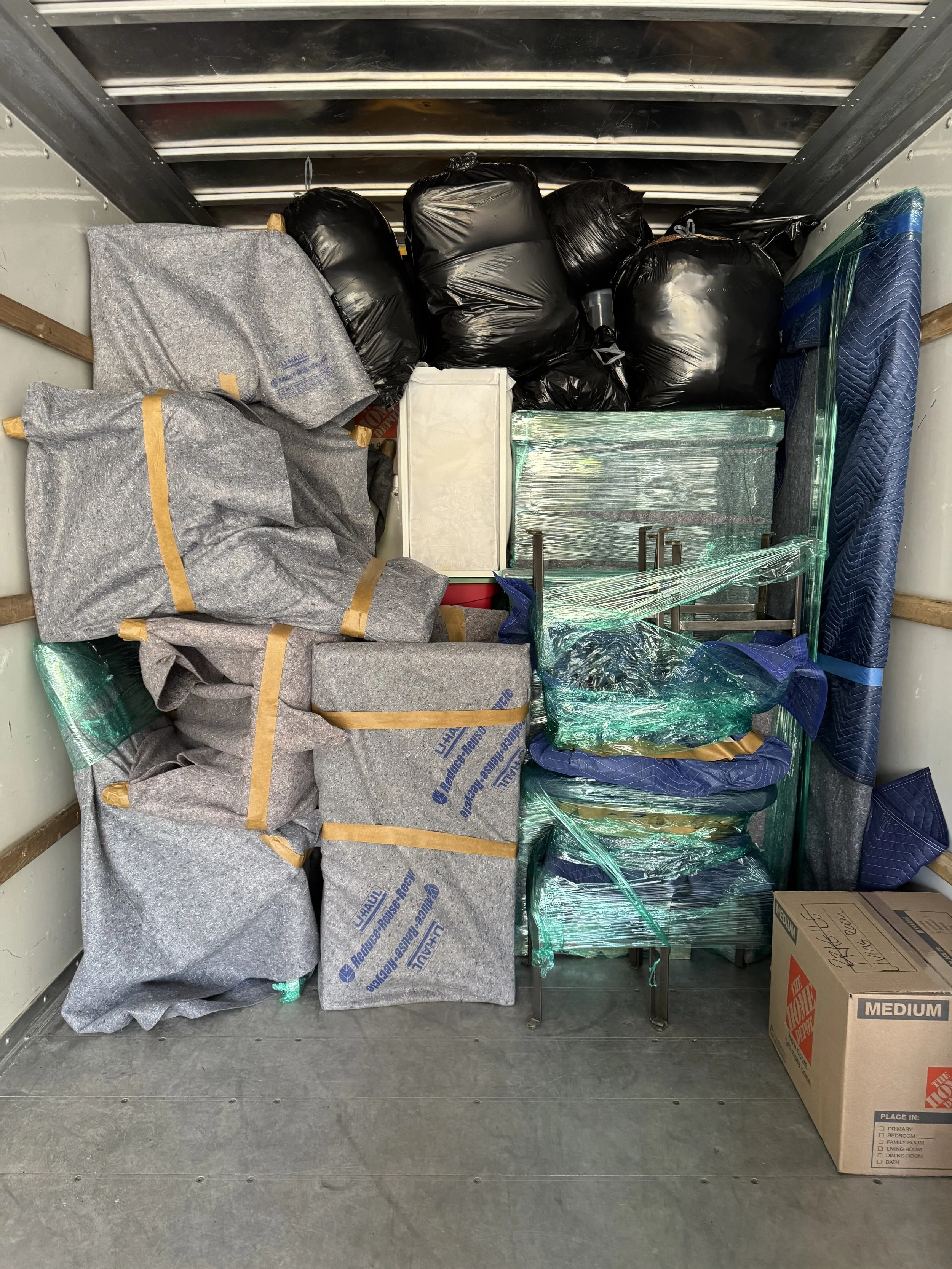 Inside a moving truck filled with wrapped furniture, black plastic bags, and a cardboard box.