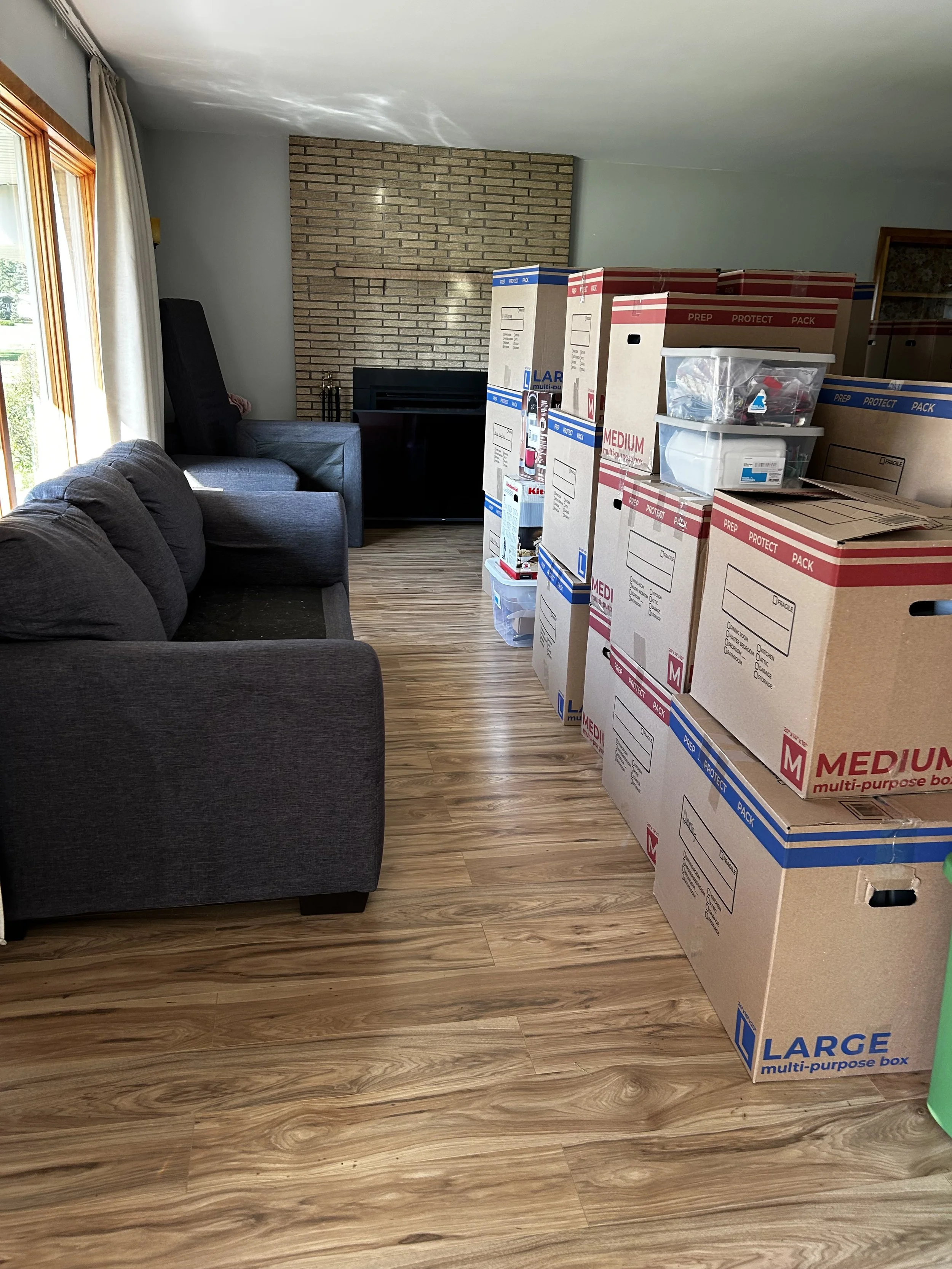 Living room with brown hardwood flooring, gray sofas, a brick fireplace, and multiple moving boxes stacked near the wall.