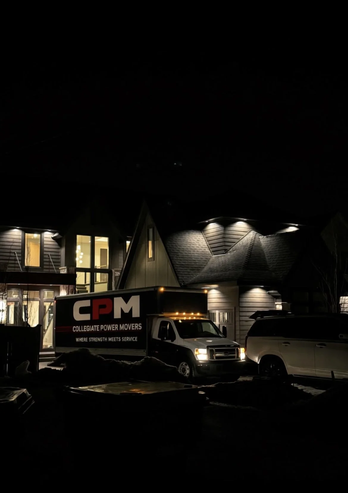 Nighttime view of a house with large windows illuminated from inside, a parked truck with the logo "CPM Collegiate Power Movers" on the side, and another vehicle nearby.
