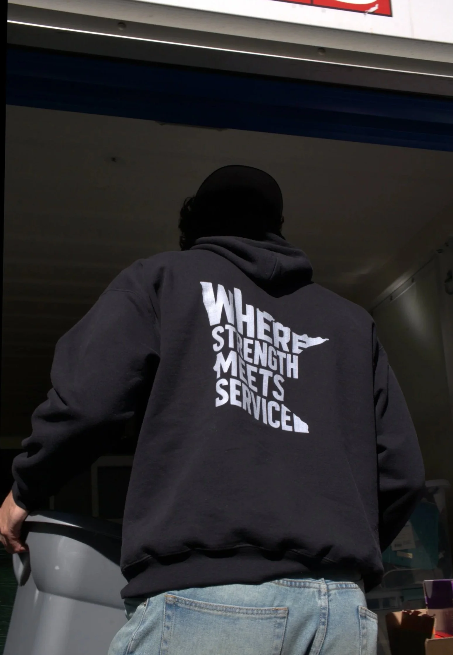 A person wearing a black hoodie with white text on the back that reads, 'WHERE STRENGTH MEETS SERVICE', standing in front of an ATM at a gas station or convenience store.