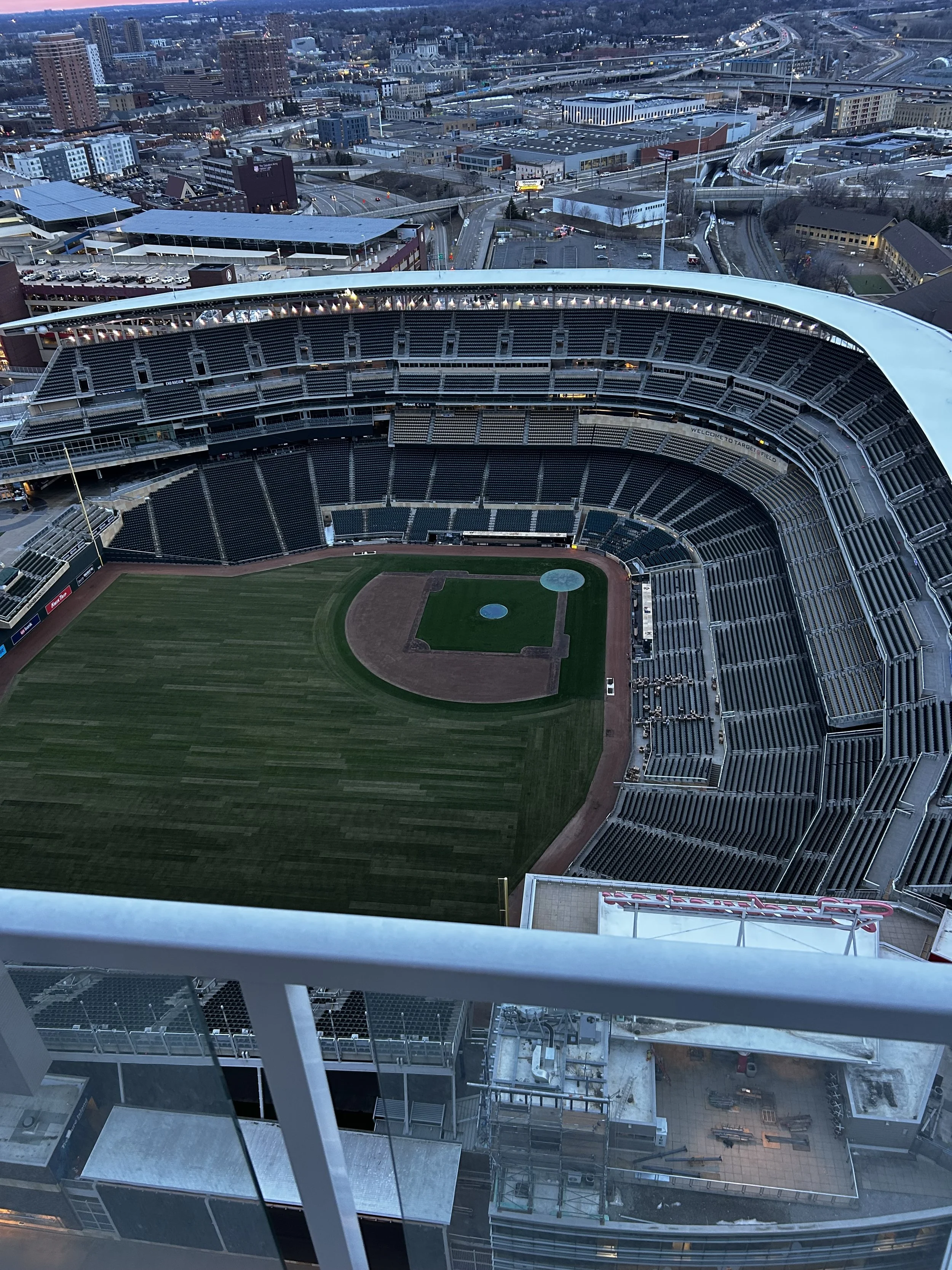 Aerial view of a baseball stadium with empty seats and a baseball field, surrounded by an urban cityscape with roads and buildings.