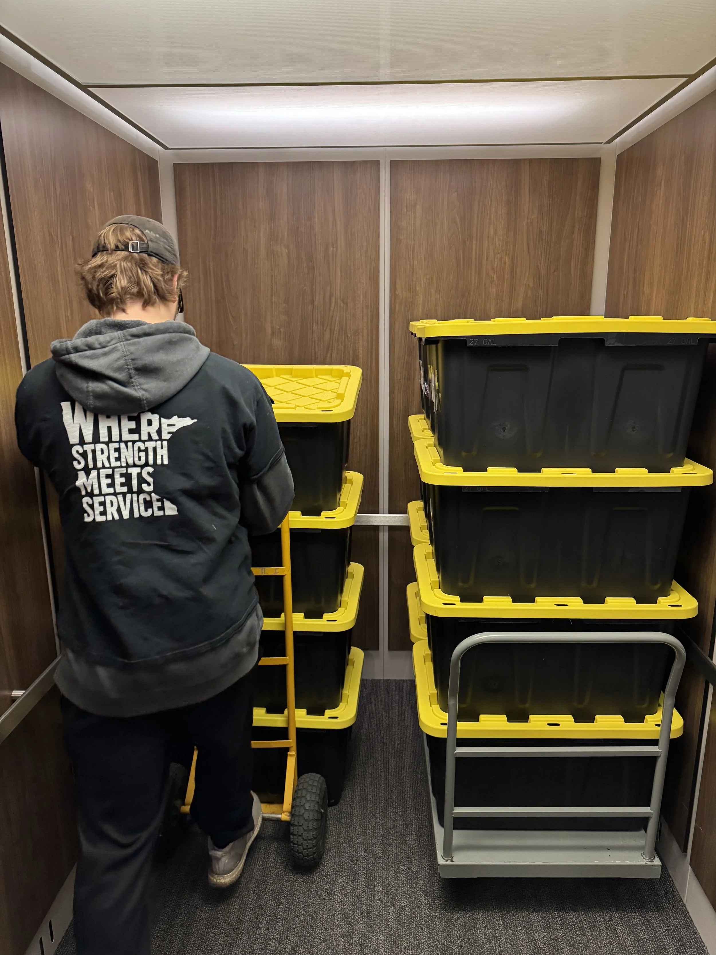 A person with a black hoodie that has the words "WHERE STRENGTH MEETS SERVICE" on the back, standing in an elevator with wooden-paneled walls, pushing a yellow hand truck loaded with black plastic storage bins with yellow lids, and another set of similar stacked bins beside it.