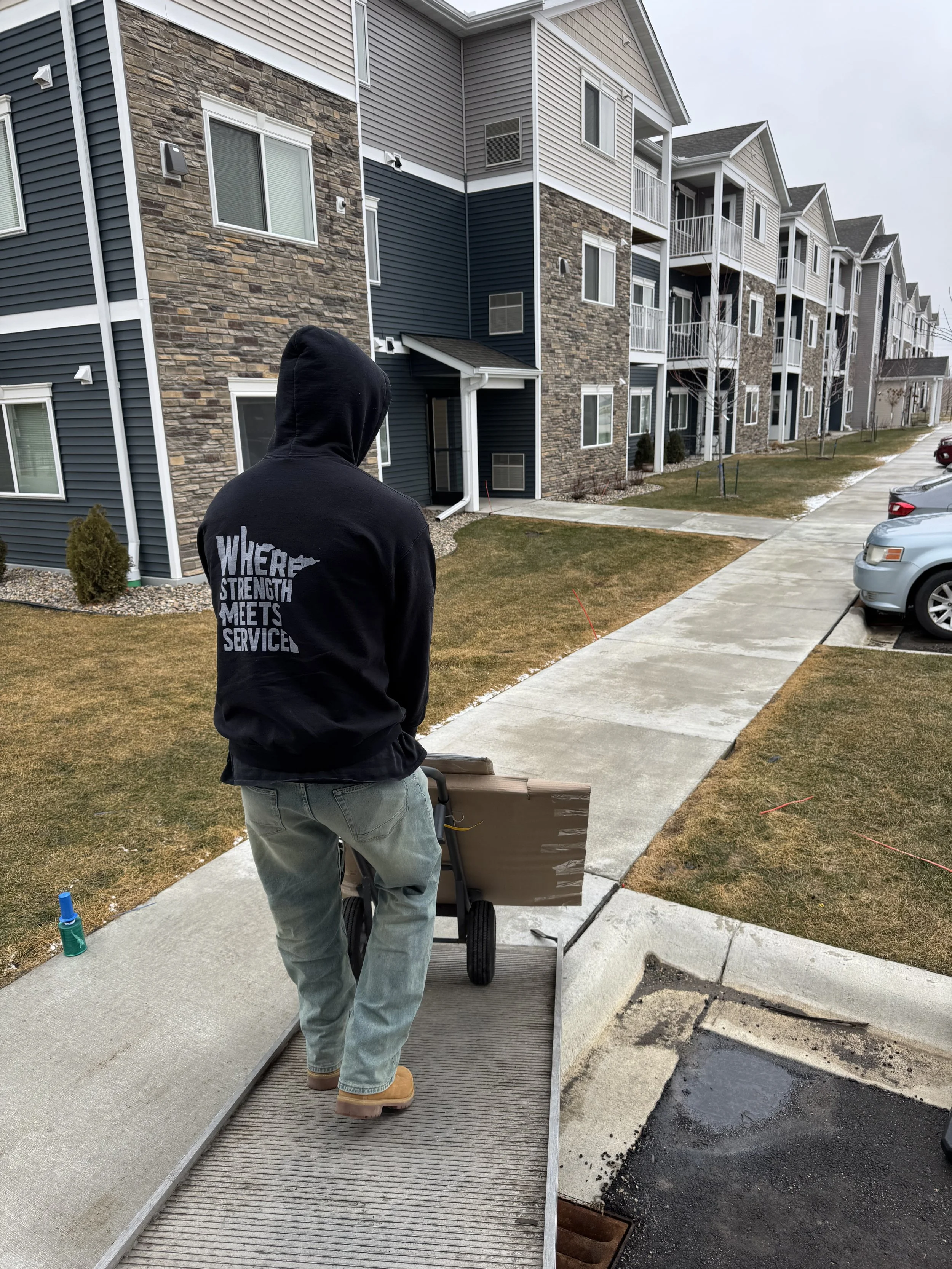 A person wearing a black hoodie with the words 'WHERE STRENGTH MEETS SERVICE' on the back walks with a hand truck loaded with a flat package along a concrete sidewalk outside a multi-family apartment complex.