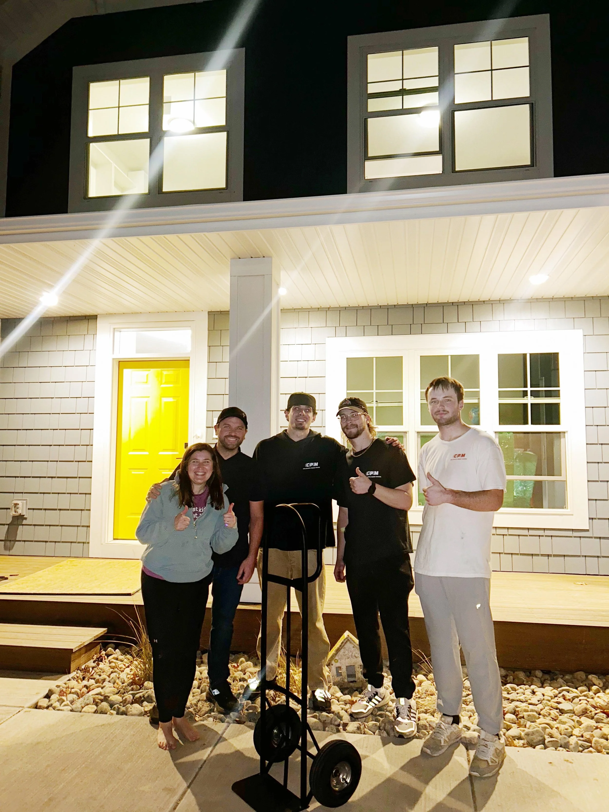 A group of five people standing outside a house at night, smiling and posing for a photo. The house has grey shingles, a bright yellow door, and white trim around the windows. The group appears happy, with some giving thumbs-up or showing a peace sign. The house has a porch with a deck, and the group is standing on a paved walkway with decorative rocks.