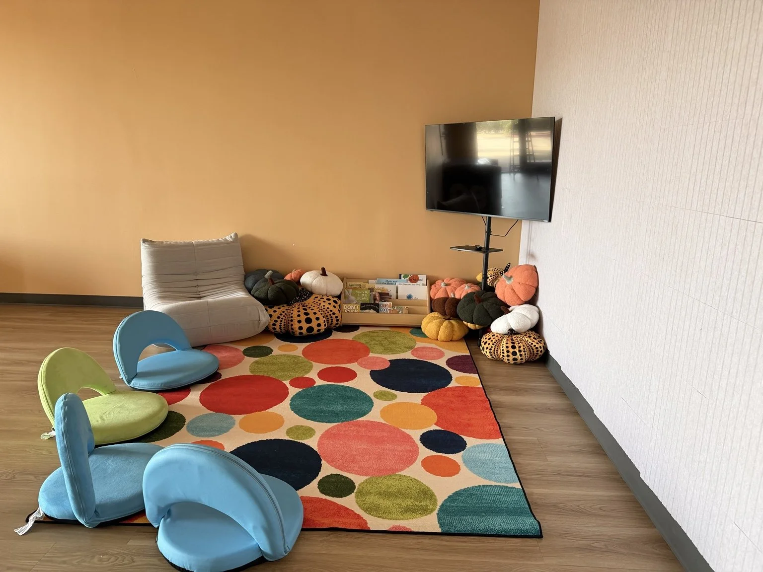 A children's reading and play area with a colorful polka dot rug, plush pumpkin and pumpkin-shaped cushions, a cozy gray chair, three small blue and green chairs, and a mounted flat-screen TV on a stand.