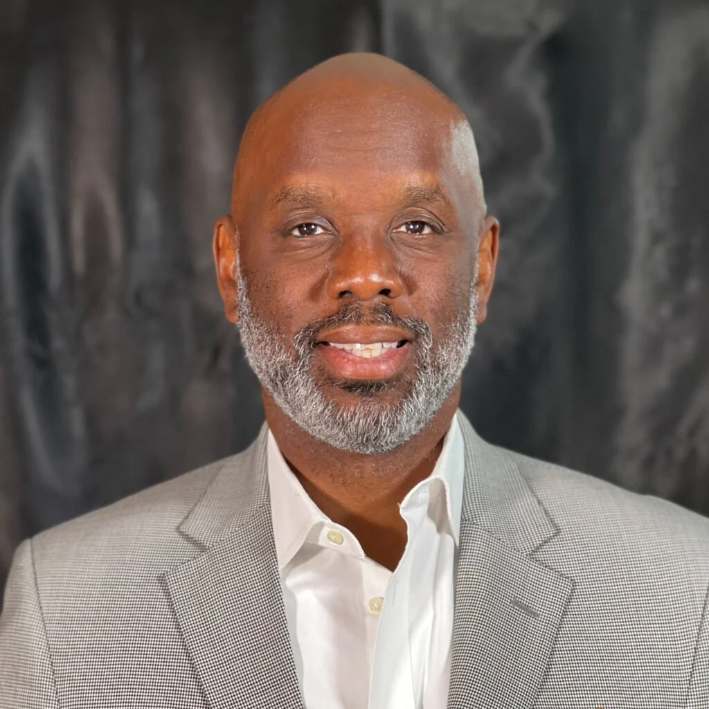 A middle-aged African American man with a gray beard, dressed in a checked gray blazer and white shirt, posing in front of a dark, textured background.