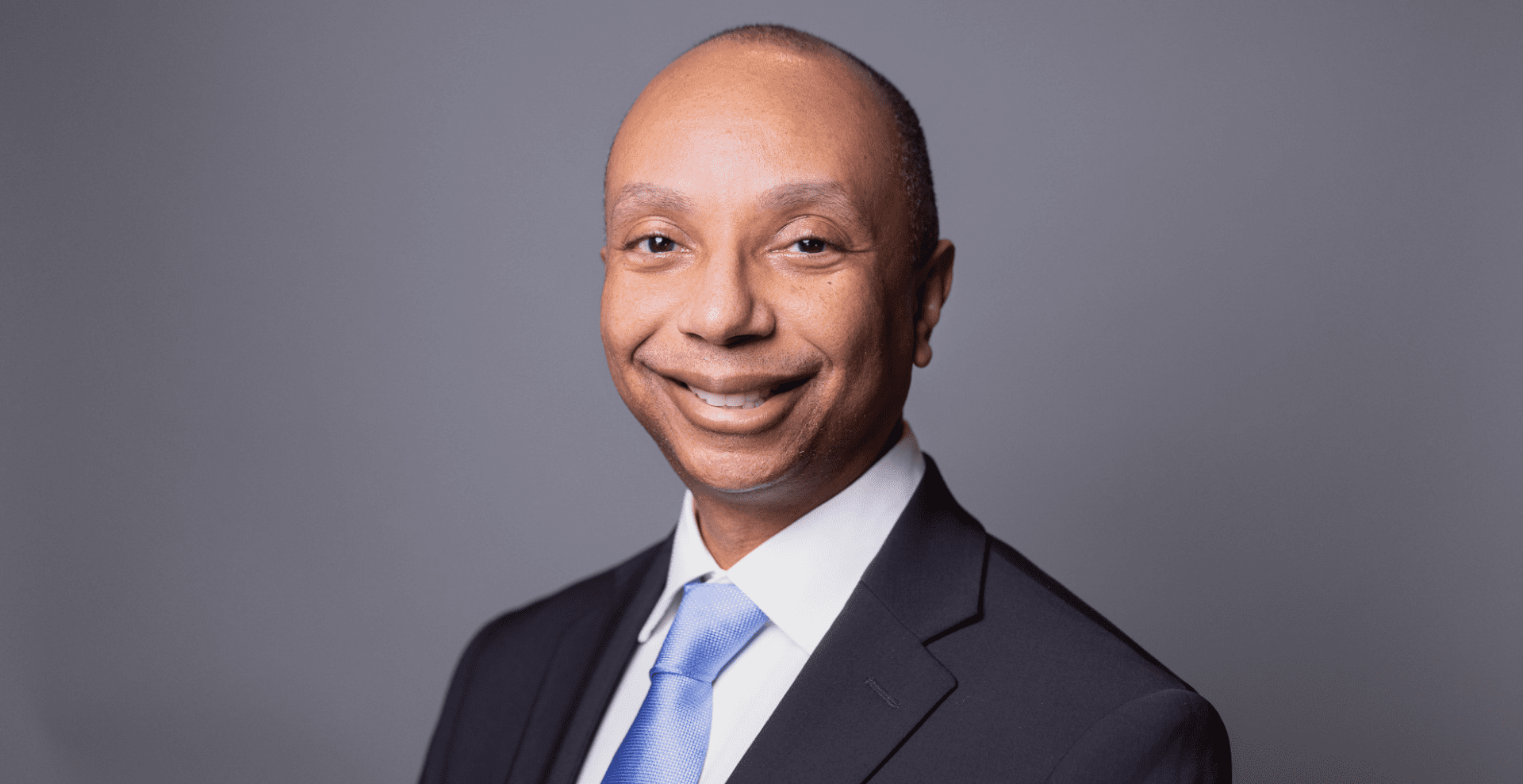 Headshot of a smiling middle-aged African American man in a dark suit, white shirt, and blue tie, against a gray background.