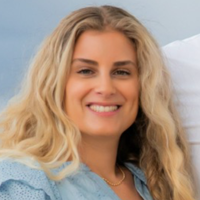 A woman with long blonde curly hair smiling, wearing a light blue top, with a white pillow behind her.