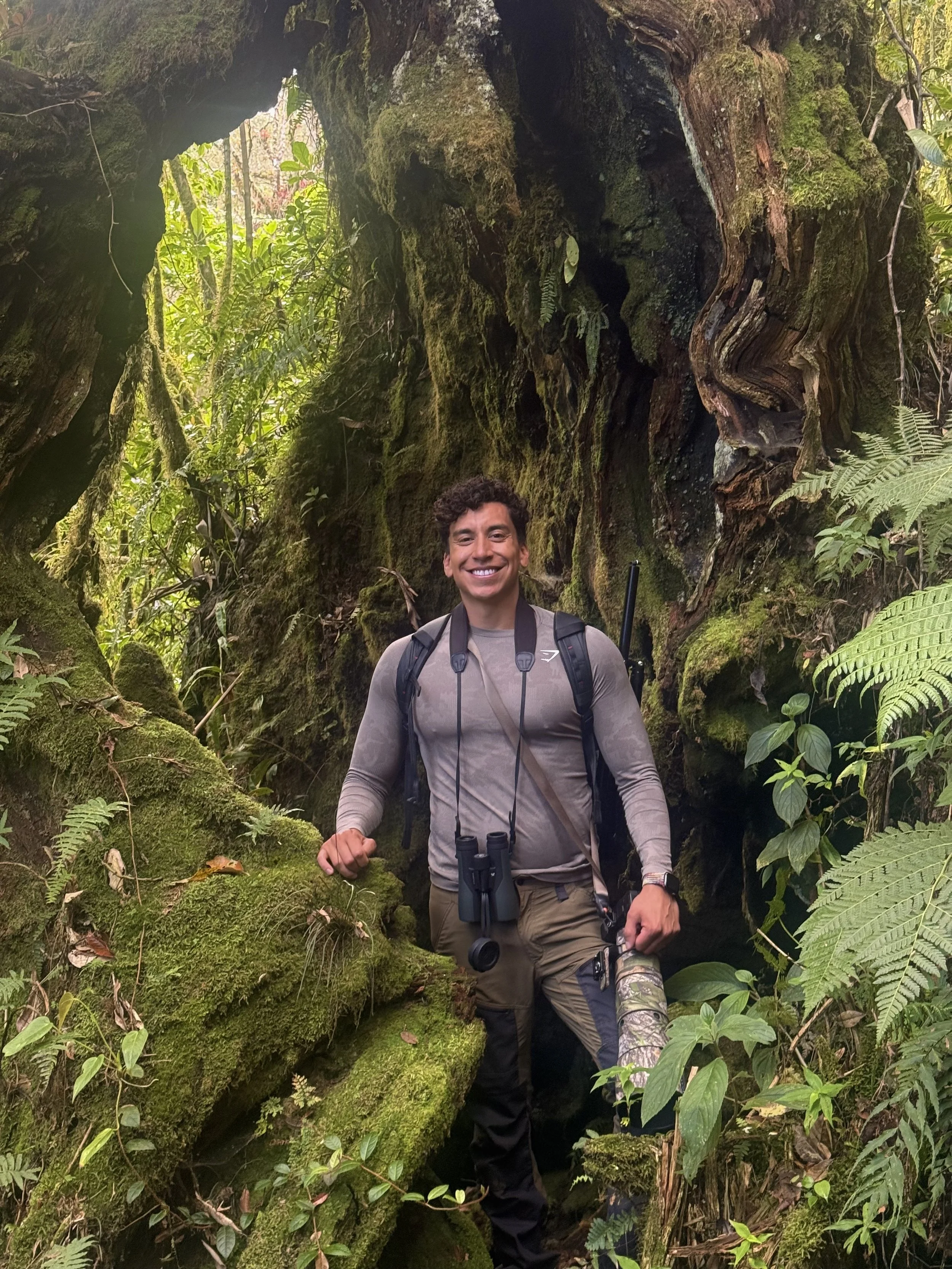 A man with a backpack and binoculars standing in a lush, moss-covered forest, smiling at the camera.
