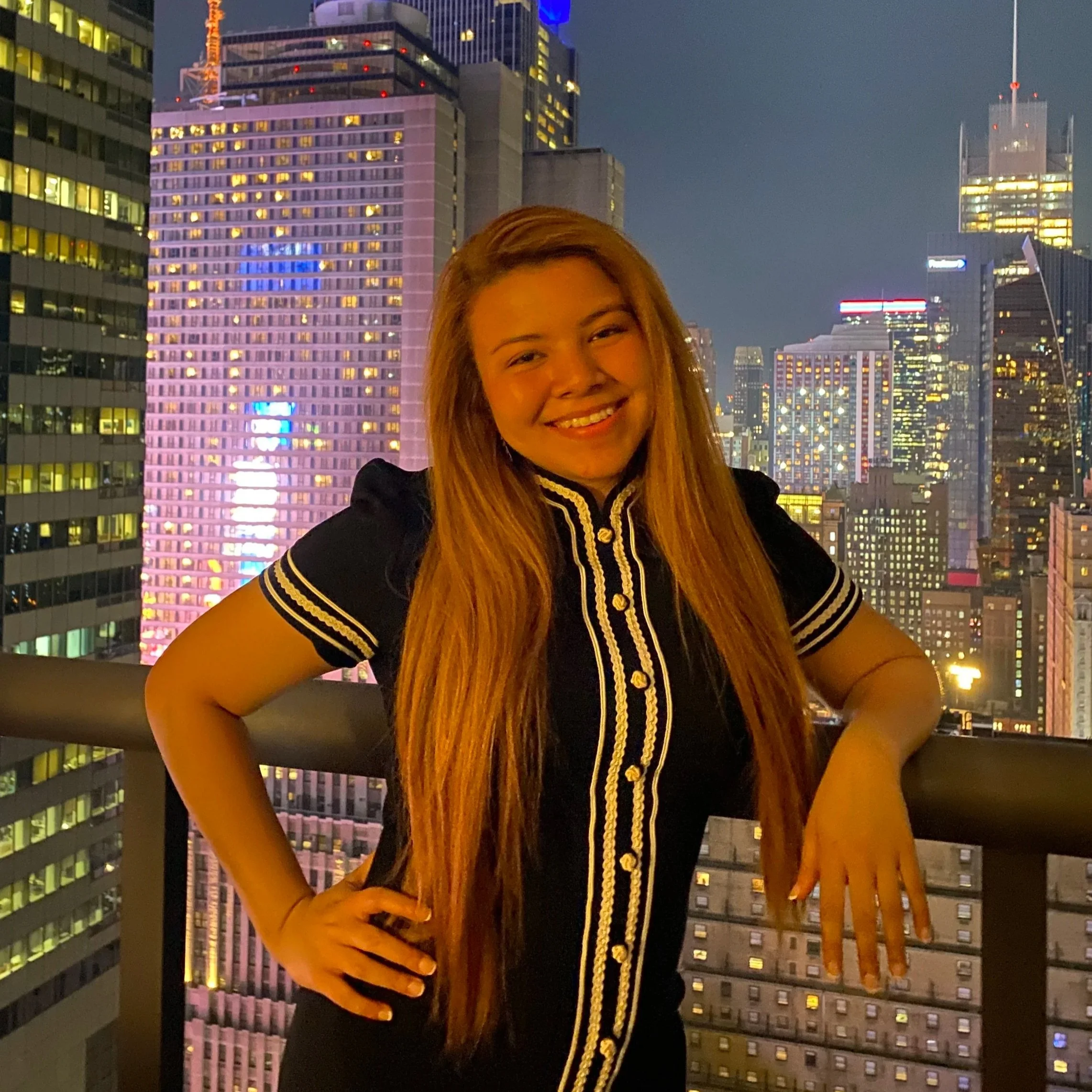 A smiling young woman with long hair stands on a balcony with a city skyline at night in the background. The city is lit up with numerous windows and colorful lights.