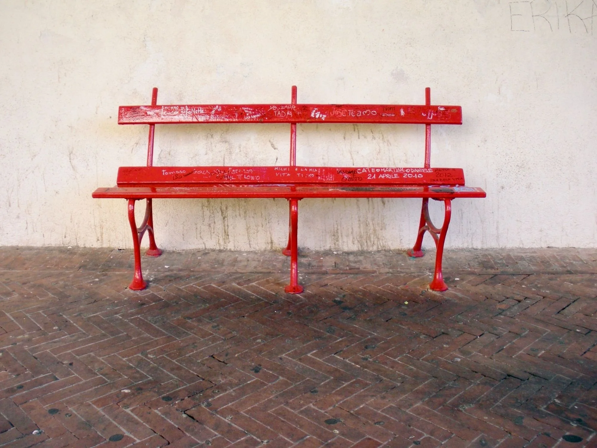 Red graffiti-covered bench against a weathered wall in Udine, Italy.