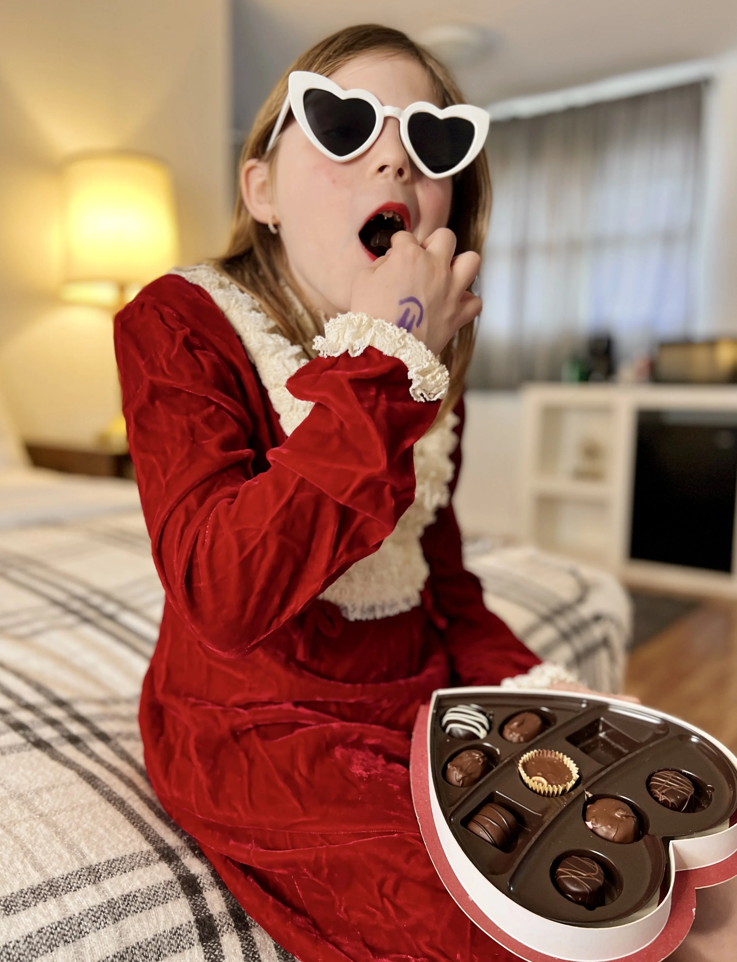 Girl in a red velvet dress and heart-shaped sunglasses eating chocolates at Dame Fortune’s Cottage Court.