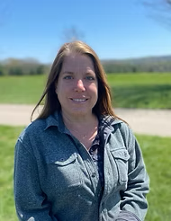 Woman with long brown hair smiling outdoors in front of green fields and blue sky