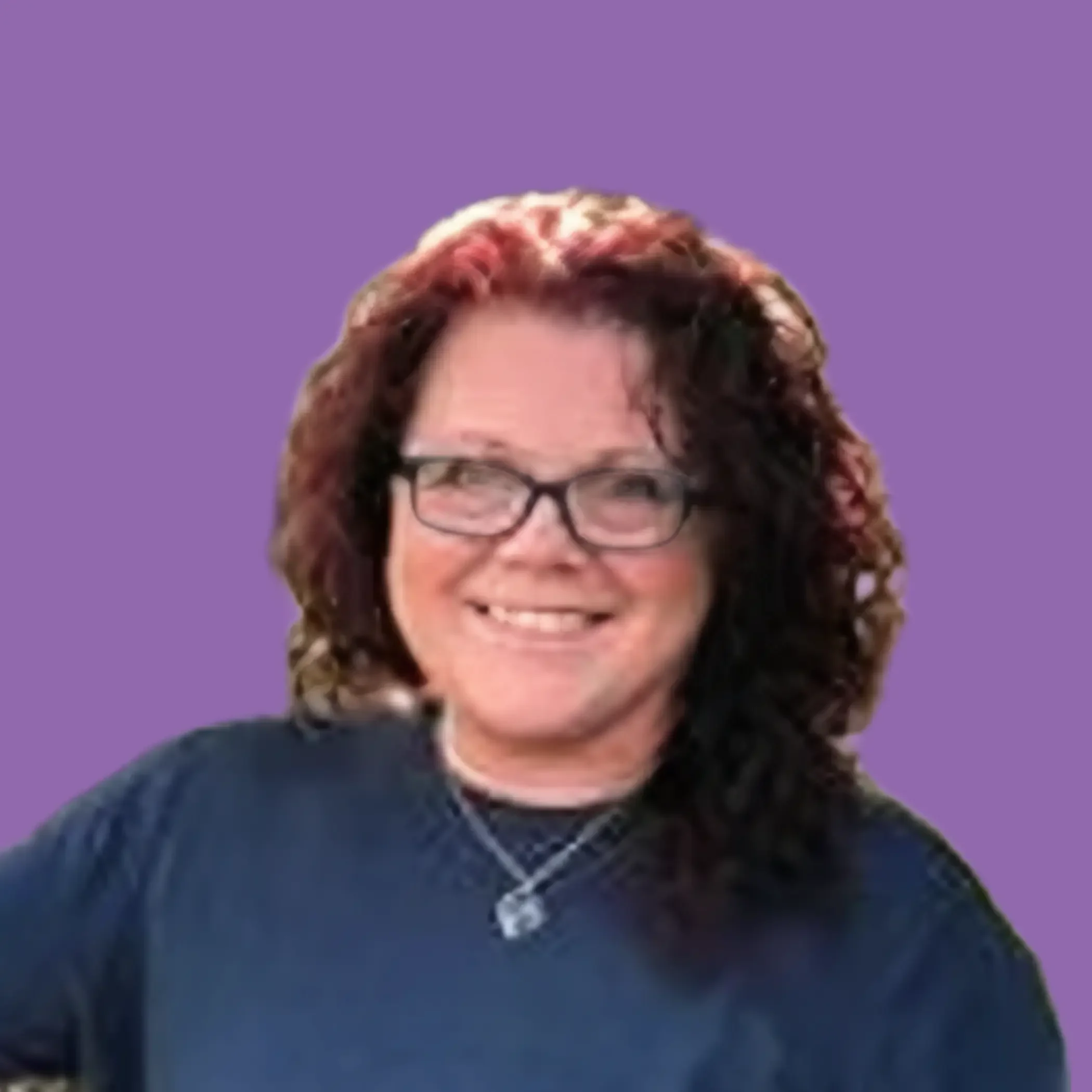A woman with curly red hair and glasses smiling outdoors, wearing a blue long-sleeve shirt, standing next to a tree.