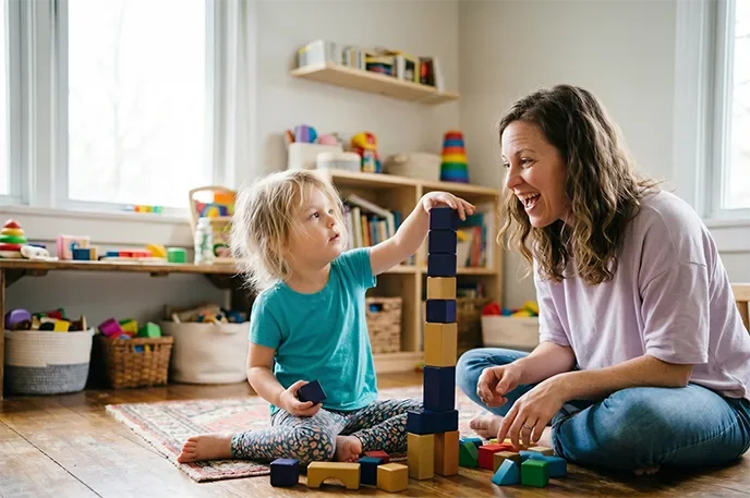 A young girl and an adult woman playing with wooden building blocks on the floor of a brightly lit room.