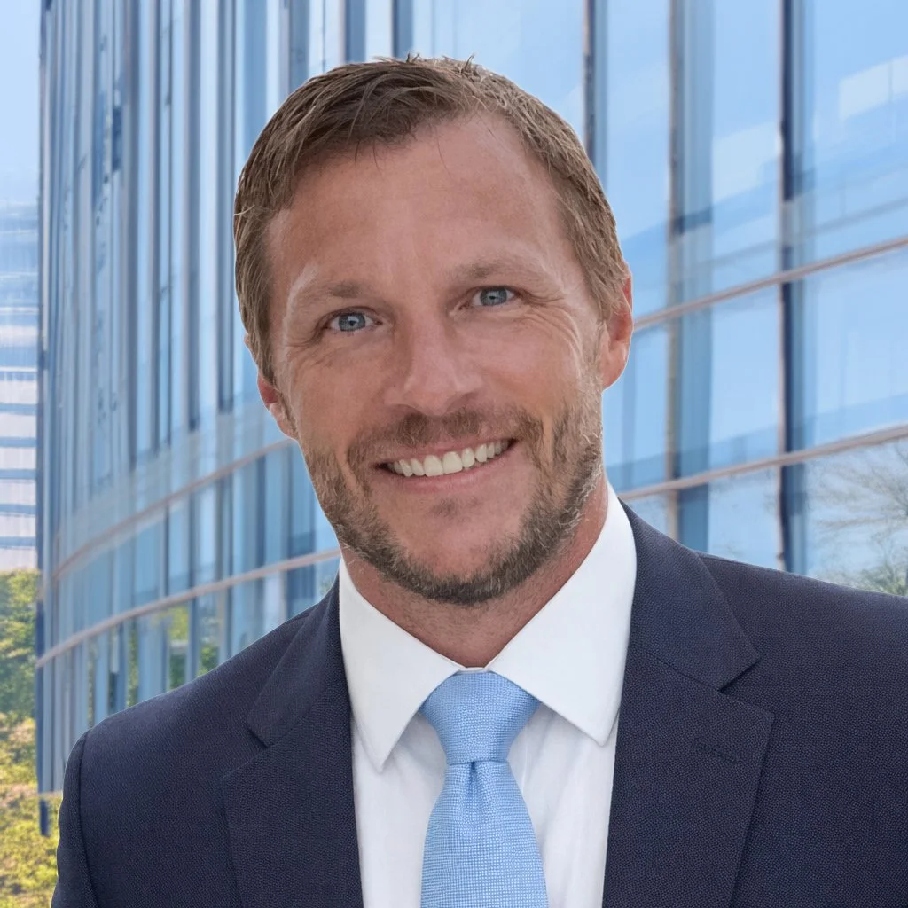 A smiling man in a suit and light blue tie standing in front of a modern glass building.