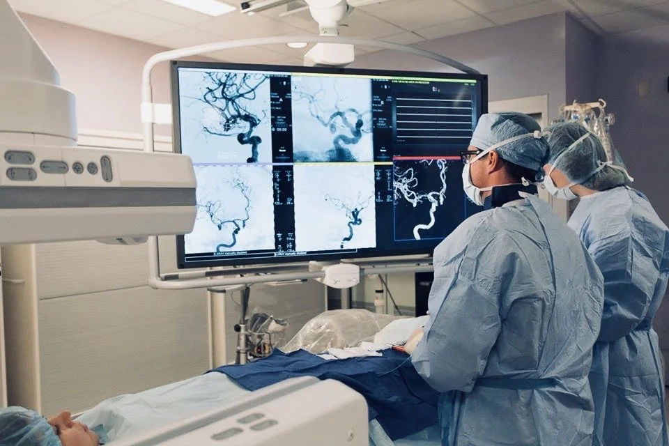 Surgeons in an operating room examining brain angiogram images on a large screen during surgery, with medical equipment and a patient on the operating table.