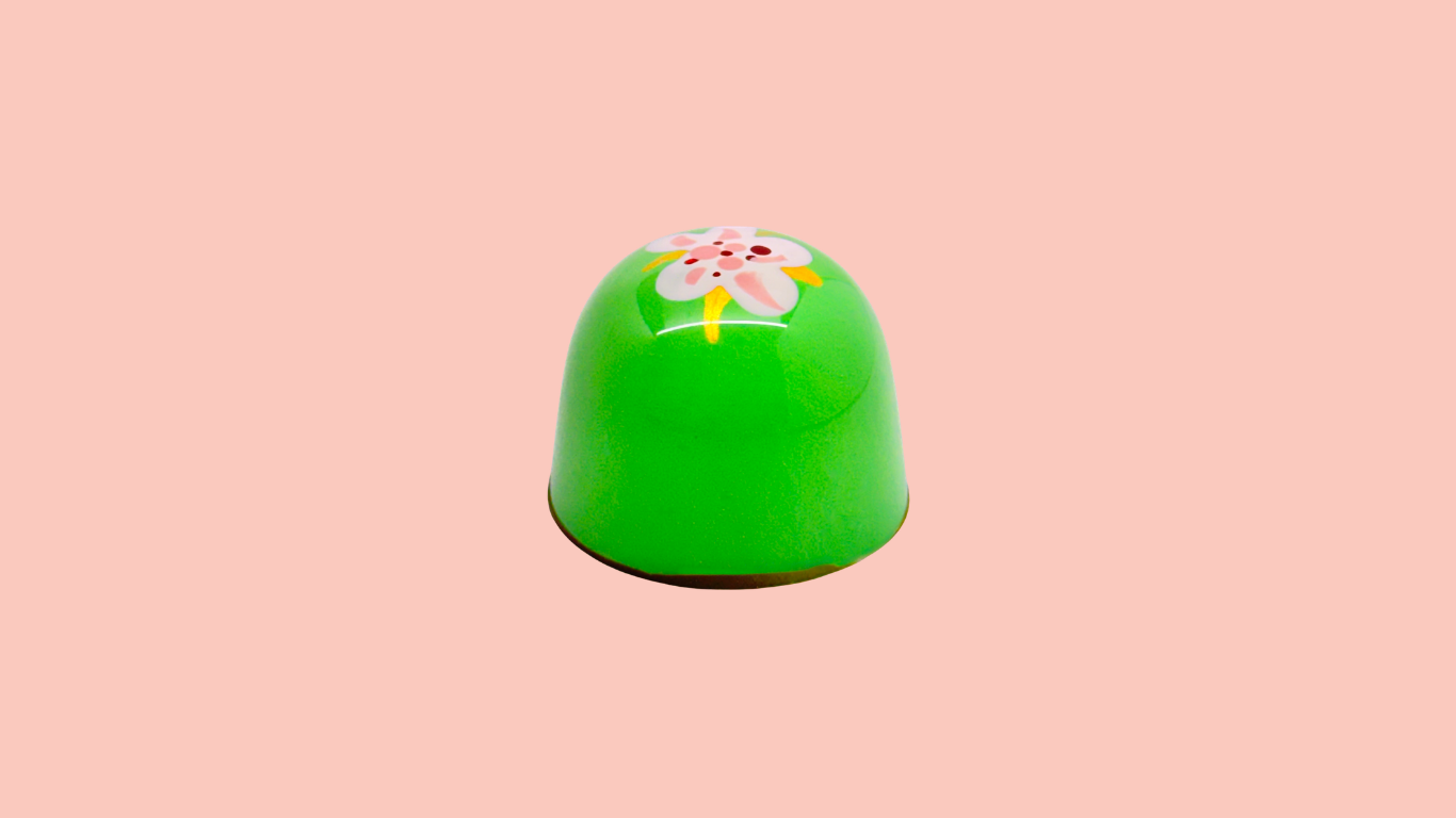 A green, rounded chocolate candy with a pink floral pattern on top, placed against a light pink background.