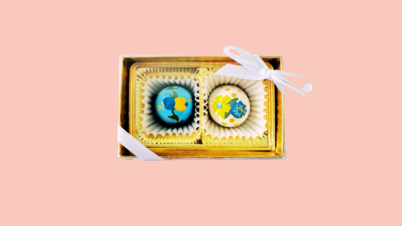 A gift box with two round decorated chocolates inside, each in individual paper cups, tied with a white ribbon on a pink background.
