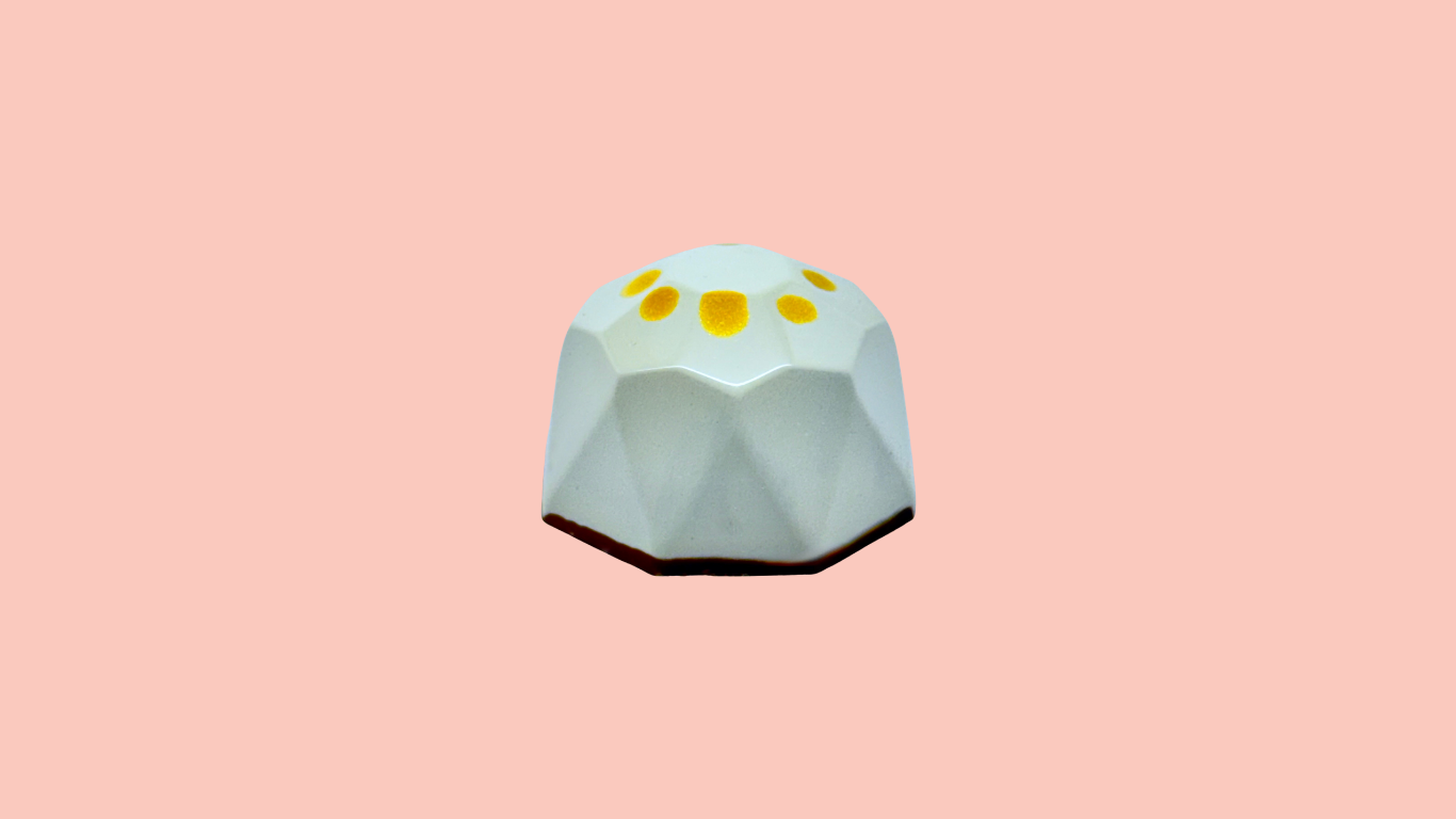 A white, geometric-shaped bonbon with yellow dots on top, set against a pink background.