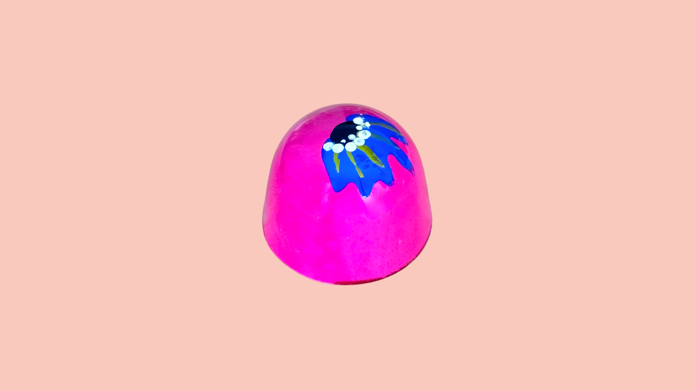 A pink, rounded object with a painted blue and green leafy design on top, set against a light pink background.