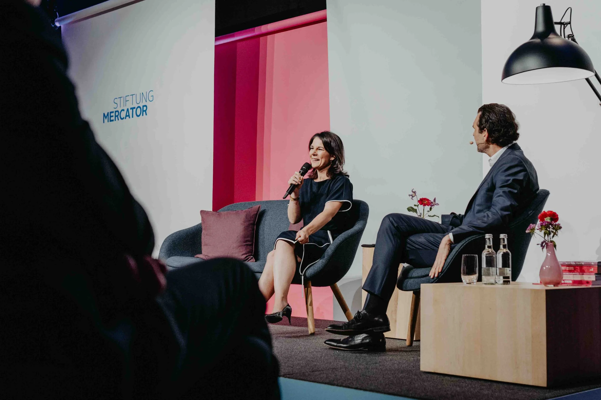 Zwei Personen im Gespräch auf einer Bühne bei einer Podiumsdiskussion vor pinkfarbenem Bühnenaufbau mit Stiftung-Mercator-Logo